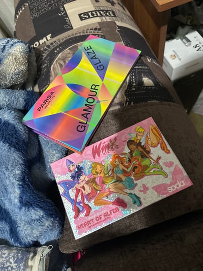 Two eyeshadow palettes are displayed on a soft, textured surface, possibly a blanket or upholstery. The palette on the left has a holographic, iridescent cover with geometric patterns in shades of yellow, green, blue, and pink. Text on this palette reads "GLAZE" and "GLAMOUR," with "PARISA cosmetics" below. The palette on the right features an illustration of the characters from the animated series Winx Club on a pink and silver background. Text on this palette includes "soda," "Wink," and "HEART OF ALFEA eyeshadow palette #magicalpower." In the background, a white box with "A8" and "P PATRIOT" is visible, along with other indistinct items. The lighting suggests it is indoors, possibly in a home environment. The overall scene is casual and domestic, with the focus on the cosmetic products.
