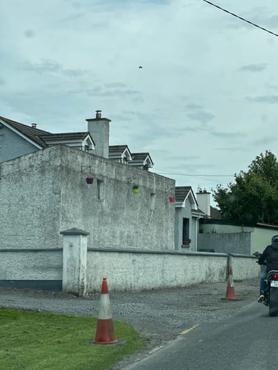 The image depicts a street scene in Carlow, Ireland, characterized by a cloudy sky and overcast conditions. A light blue house with a grey, textured stucco exterior and a dark tiled roof dominates the left side of the frame. The house features multiple dormer windows with dark frames. Adjacent to it is a lower grey wall, also textured, running horizontally across the mid-ground. Several colorful hanging flower baskets, one purple and one green, are attached to the wall at varying heights. A white concrete post stands in front of the wall. In the foreground, a grey gravel driveway borders the grass on the left. Two orange and white traffic cones are visible on the gravel and the edge of the road. On the right side of the image, a person rides a motorcycle away from the camera on an asphalt road. The motorcycle has a license plate that reads "191-C-1383". The rider is wearing dark clothing and a helmet. Behind the house and wall, lush green trees are visible on the right side of the image. A thin black wire runs diagonally across the upper portion of the frame.