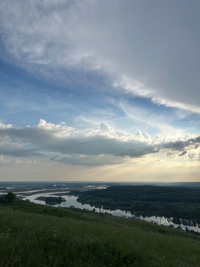 An aerial view captures a wide, meandering river winding through a lush, forested landscape. The river's surface reflects the sky, which is a dramatic blend of blue and white clouds, with rays of sunlight piercing through in places. In the foreground, a grassy hillside slopes downwards towards the river, dotted with small white wildflowers. The scene is devoid of people or any visible human activity, presenting a natural and serene panorama. The location is identified as Britvino, Russia.