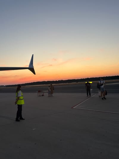 An airport tarmac at sunset shows the tail of a Ryanair airplane with its name visible. A person wearing a bright yellow safety vest and dark pants stands on the tarmac. In the background, a group of people are walking across the tarmac, some with luggage. The sky displays a vibrant orange and pink sunset with a line of dark trees in the distance. The location is the Niederrhein Airport Weeze (NRN) in Weeze, Germany.