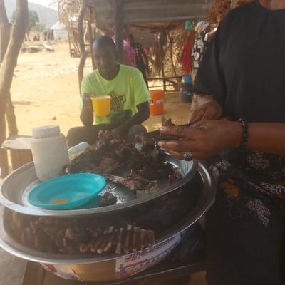 A person arranges cooked meat on a large metal platter alongside ribs. The platter holds a small turquoise plate with spice and a white container. A man in a green shirt holding a yellow cup sits in the background. The setting is outdoors, near a building with visible wooden beams.