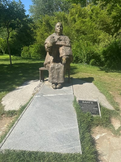 This image displays a bronze statue of the Bulgarian mystic Vanga, located outdoors in General Todorov, Bulgaria. The sculpture depicts an elderly woman seated on a low, simple stool, draped in a textured robe with her hands resting in her lap. Her facial expression is serene and contemplative.
The statue rests on a light grey granite slab that forms a narrow pathway. To the right of the statue, a black informational plaque is embedded within the granite. The plaque features white Cyrillic text, stating "ВАНГА" (Vanga), and indicates that the sculpture was created by Emil Popov on the occasion of her 100th birthday, donated by the "Vanga" Foundation.
The surrounding environment is a verdant park or garden. A dense backdrop of various green trees and bushes fills the midground and background. Lush green grass covers the ground around the granite pathway, with a lighter, possibly gravel, path running parallel to the left of the main pathway. The sky above is clear blue, visible in the upper portion of the image. The scene is illuminated by bright natural sunlight, casting distinct shadows from the statue and surrounding foliage onto the ground.