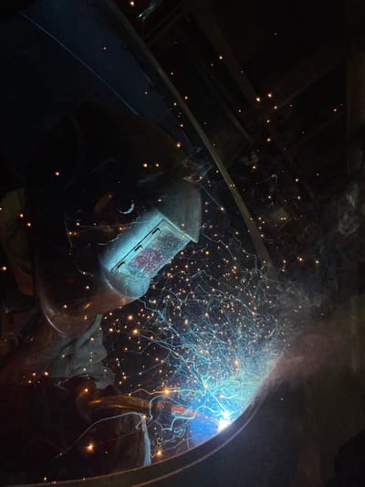 A person wearing a welding helmet is welding metal in a dark, enclosed space. Sparks are flying from the point of welding, illuminating the area around the work. The location is unknown, there is no information available to place it in Tomba, Montenegro.