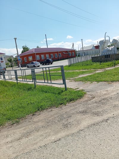 The image depicts a sunny day in Otradnaya, Russia. In the background, a two-story red brick building with a maroon roof stands prominently. Several windows are visible on the building's facade. To the right of the building, a modern sculpture of two mammoths is present, likely a local landmark. A metal fence separates the building and sculpture from a grassy area in the foreground. Several vehicles, including a silver sedan and a black SUV, are parked or driving on the road in front of the building. The scene includes a utility pole with wires overhead and a blue bus stop sign. The foreground is dominated by a dirt and gravel road and a well-maintained grassy area.