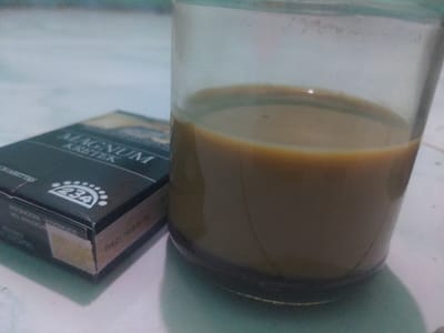 The image shows a glass of coffee, partially filled, resting on a white surface. Next to the glass, a pack of "Magnum Kretek" cigarettes, black with silver lettering, is partially visible. The cigarette pack bears a warning sign in Indonesian and has the number "234" prominently displayed. The background shows a blurry, light blue surface. The image was taken in Batam, Indonesia, at the coordinates 1.047 latitude and 103.944 longitude. The scene is presented in a simple, unassuming manner, likely captured indoors on a table or countertop.