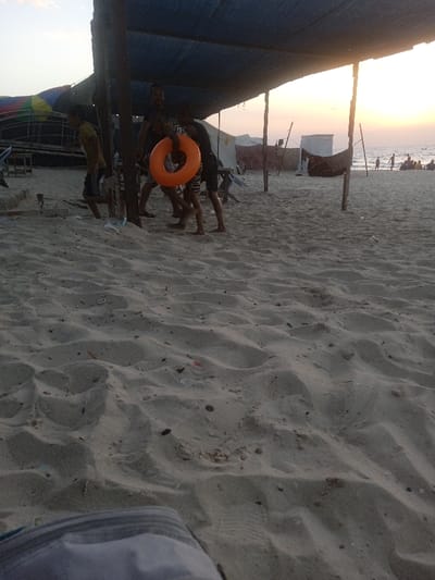The image depicts a sandy beach scene in Al-Mawasi, Palestinian Territories, during either sunset or sunrise, evidenced by the warm glow on the horizon. In the midground, a makeshift canopy, constructed from a blue tarp supported by wooden poles, shades an area of the beach. Under this canopy, at least four individuals are visible, mostly male and barefoot. One person, wearing a dark shirt and patterned shorts, holds a prominent orange inflatable swimming ring. Another individual in a yellow shirt is partially visible to the left, walking away from the center. The figures are partially silhouetted against the brighter background light.
The foreground consists of undisturbed beach sand, showing undulations and scattered small debris. In the background, beyond the canopy, temporary structures including white tent-like shelters and wooden frames are present. Further back, near the water's edge, several indistinct figures are visible, some standing and some seated, indicative of people gathered on the beach. A dark object, possibly a small boat or debris, is near the background structures closer to the water. The sky transitions from a pale blue at the top to a bright orange-yellow near the horizon, reflecting the low sun. The lower-left corner of the image shows a blurred portion of a grey fabric object.