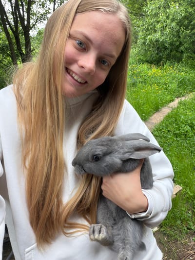A young person with long, blonde hair is pictured smiling broadly at the camera while gently cradling a small, fluffy grey rabbit. The image is taken from a close perspective, likely a selfie, with the subjects appearing to be outdoors and tilted, possibly lying down or holding the camera at an angle.

The person is wearing a white long-sleeved top or hoodie, and a white Apple Watch is visible on their left wrist. Their expression conveys warmth and happiness, with bright blue eyes and a wide smile. The grey rabbit, featuring dark, alert eyes, is nestled comfortably against the person's chest and hair, appearing calm and at ease.

The background is a vibrant, lush green landscape filled with various trees and grassy areas. Scattered among the grass are small yellow wildflowers, suggesting a pleasant outdoor setting like a garden, park, or a natural woodland edge. A faint wooden or concrete border can be seen in the distant mid-ground, implying a pathway or a defined area within the greenery. The lighting indicates it is daytime, with ample natural light illuminating the scene. The overall mood is peaceful and affectionate, highlighting a tender moment between the person and the animal. There is no visible text in the image.