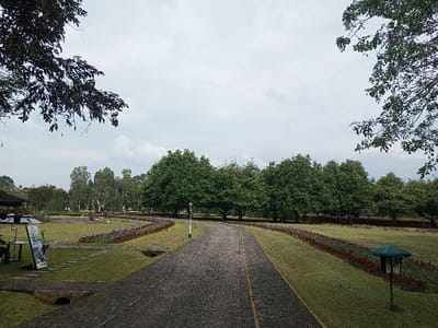 A park scene under a cloudy sky, showcasing a cobblestone path winding between green lawns. Rows of red flowers border the path. Tall, leafy trees line the background and frame the top corners of the image. A small structure with a booth sits on the left, while a decorative lamp post stands on the right, adding visual interest to the landscape.
