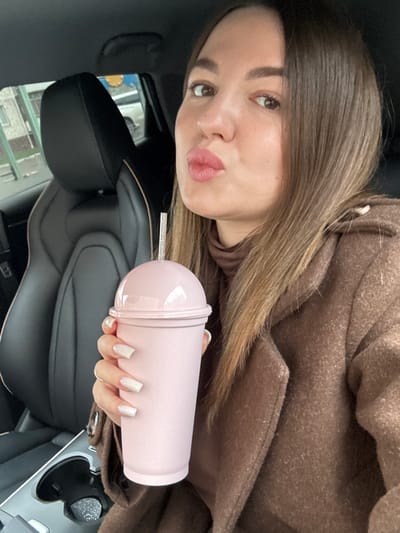 A woman is in the driver's seat of a car, holding a pink cup with a straw. She is wearing a brown coat and has brown hair. The car is likely parked in Likino-Dulyovo, Russia, based on street sign visible in the reflection on the windshield.