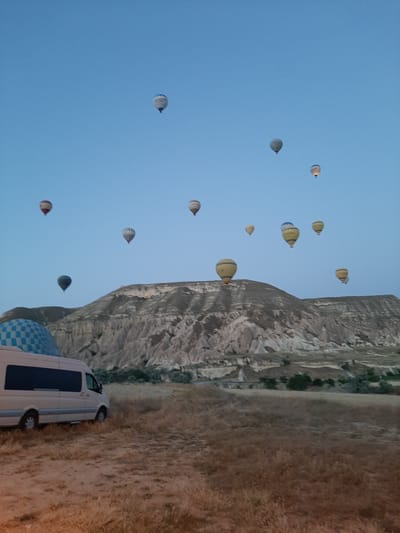 In Avanos, Turkey, a fleet of hot air balloons fills the pale blue pre-dawn sky over a rugged, mountainous landscape. The balloons, in various colors and patterns, are ascending or already airborne. In the foreground, a light-colored minivan is parked on a dirt and dry grass field. The terrain consists of dry vegetation and sandy soil, with a distinctive layered rock formation forming the backdrop. A blue and white checkered dome, possibly part of a structure, is visible on the left side of the image. The scene suggests an early morning hot air balloon launch, a common tourist activity in Cappadocia.
