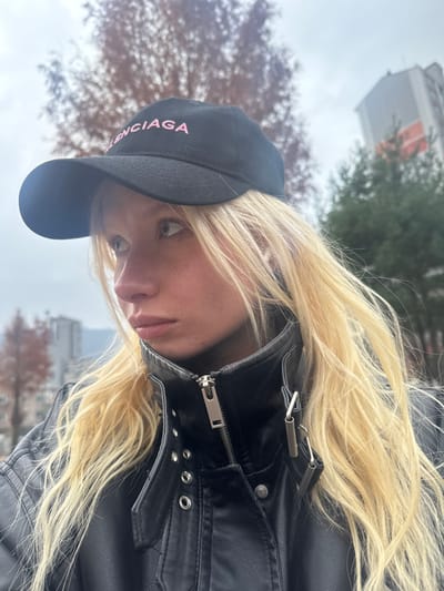 A woman with blonde hair and light eyes peers at the camera. She wears a black baseball cap with "Balenciaga" in pink lettering and a black leather jacket with silver hardware. The background shows a cloudy sky, bare branches of trees, and buildings. The photo is taken from a low angle, making the woman appear to be looking down.