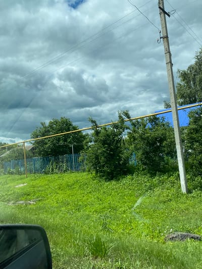 The image depicts a cloudy day in Nadezhdino, Russia. A concrete utility pole with electrical wires stands on the right side of the frame, next to a dense green area. Behind the greenery, a blue metal roof of a building is visible, along with a yellow pipe. A blue picket fence runs horizontally across the mid-ground, separating the grassy area from the trees and houses behind it. The foreground shows a rearview mirror of a vehicle, indicating the photo was taken from inside a car. The overall scene suggests a rural or suburban setting with elements of infrastructure and residential structures.
