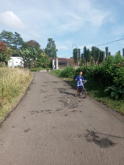 A young boy in a blue shirt and shorts stands astride a bicycle on a cracked asphalt road. Tall grass lines the left side of the road, while dense green bushes grow on the right. In the background, there are houses with fences and trees, including banana trees. The sky is blue with scattered clouds. The location is likely a rural or suburban area in Ein Chay Hnit Se, Myanmar, suggested by the combination of vegetation and simple residential structures.