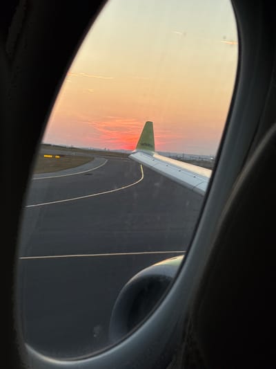 An airplane wing with the AirBaltic logo is visible through a plane window, with a sunset over the runway at Vienna International Airport (VIE) in Fischamend, Austria. The sun is low on the horizon, casting a warm orange and pink glow across the sky and clouds. The runway is dark asphalt, with illuminated markings and signs indicating its direction and boundaries. The wing is white and grey, with a green tip. The interior of the airplane window frame is dark and blurry, with the curvature of the window visible. The airport environment is visible in the distance, with low-rise buildings and infrastructure. The image captures a moment of departure or arrival during a beautiful sunset.