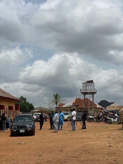 The image depicts an outdoor scene in Kassa, Nigeria, with a group of people gathered in a dirt-covered area. The sky is overcast with dramatic, dark clouds. Several buildings are visible in the background, including one with a red-tiled roof and another with a corrugated metal roof. A prominent feature is a tall, industrial-looking water tower.

In the foreground, a black Volkswagen Golf car is parked, facing the camera, with a Nigerian license plate reading "PKN 631KM." Several individuals are standing around the car and in the open area. Some are dressed in traditional Nigerian attire, while others wear casual clothing. One person is holding a camera on a tripod, suggesting a filming or photography event. Motorcycles are parked in the middle ground. The overall atmosphere appears to be a gathering or event in progress.