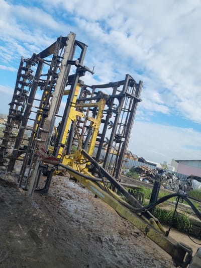 The image shows a piece of agricultural machinery, likely a cultivator or harrow, sitting on muddy ground. The machinery is composed of a heavy metal frame with multiple rows of tines or teeth designed to break up soil. The frame is painted in a combination of black and yellow. The surrounding environment appears to be a storage area or yard, possibly in an industrial or agricultural area. In the background, there is a pile of scrap metal and what seems to be a building with a corrugated metal roof. In the background a blue sky with white clouds is visible. The location is possibly Ovcharovo, Bulgaria, based on the prompt. No people are visible in the image.