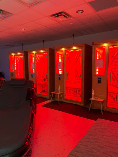 The image depicts the interior of what appears to be a wellness center in Palm Valley, United States of America, specifically a room with multiple infrared saunas. Four wooden saunas, illuminated by bright red infrared light, are lined up against a wall. Each sauna has a glass door with a handle and a digital control panel. Small wooden stools are placed outside two of the saunas, each holding a bottle of spray and a yellow cloth. In the foreground to the left, a dark recliner-like chair with a headrest is visible. The floor is a speckled white surface that reflects the red light, with a dark red carpet area in front of the saunas. The ceiling is made of acoustic tiles with fluorescent lighting fixtures, also illuminated by the red glow. The overall atmosphere is one of a modern, specialized spa or therapy facility.