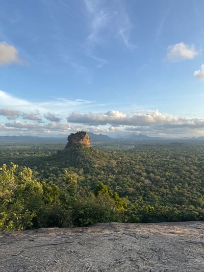 View from a rocky height in Sigiriya, Sri Lanka, overlooking a lush forest and a distant rock formation. The sky is blue with scattered clouds. A segment of rock is visible in the foreground.