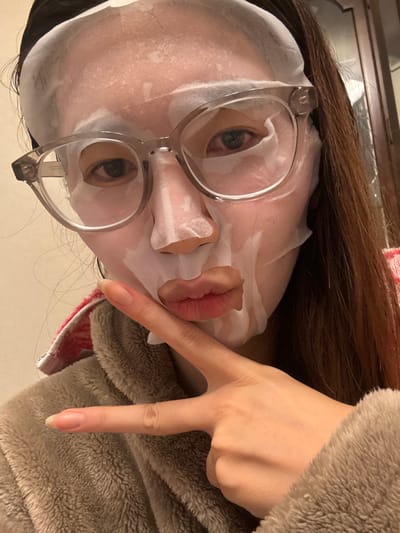 A close-up portrait depicts an individual, likely female, indoors. The subject's face is covered by a white sheet mask, featuring cutouts for the eyes, nostrils, and mouth. Transparent grey-framed eyeglasses are worn over the mask, revealing dark eyes with minor redness visible in the lower eyelids. Long, dark brown hair frames the face, pulled back with a dark headband. The mouth, visible through the mask's opening, is pursed. The individual is wearing a light brown, fluffy robe, with a red and white striped lining visible near the left shoulder. The right hand is raised, performing a 'V' or 'peace' sign gesture; fingernails are natural and unpolished. The background is an indistinct indoor environment, consisting of a light-colored wall and a darker, possibly wooden, vertical surface on the right. The image contains no visual information to confirm a specific location within Kawasaki, Japan.