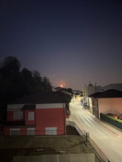 An elevated, nighttime view of a street in Balerna, Switzerland. The street is paved and lined with buildings on both sides. The sky is a dark blue, with a faint glow on the horizon and a bright object, likely the moon, visible in the upper left quadrant of the scene. Several cars are parked along the street, and a few are in motion, indicated by light trails. There are trees on a hillside to the left of the street, and a red building with white-trimmed windows is prominent in the foreground on the left. The overall scene depicts a quiet, urban street at night, illuminated by artificial light and the moon.