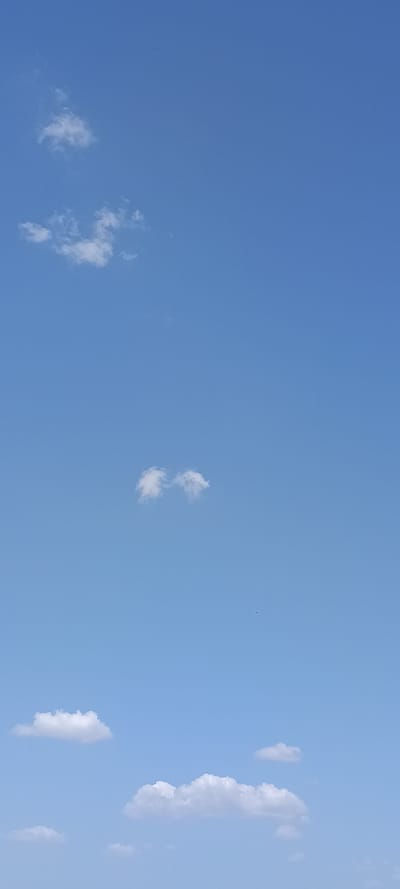 This media file depicts a bright blue sky with scattered, fluffy white clouds. The image is oriented vertically, with the sky filling the entire frame. Several small clouds are visible, with a larger, more prominent cloud formation on the far right side of the image. The sky appears clear and vast, suggesting good weather. The lighting indicates it is daytime, likely late morning or afternoon, given the brightness. There are no people, objects, or text visible in the image. The scene is a natural outdoor setting, specifically a sky view. The image conveys a sense of openness and tranquility. The location context provided is Vilseck, Germany, but this cannot be visually confirmed from the image itself.