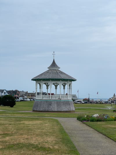 The image shows a picturesque scene in Oak Bluffs, USA. The primary subject is a charming white gazebo with green trim and a grey, multi-sided roof, topped with a weather vane. The gazebo's intricate architectural details, including arched openings and decorative railings, suggest it is a historical structure.
Adjacent to the gazebo is a well-maintained park with lush green grass, a gravel path, and a flower bed bursting with colorful blooms. In the background, a line of quaint houses with varying architectural styles and colors adds to the scene's charm. Beyond the houses, a street is visible with parked cars, indicating a populated area. American flags on poles fly in the distance, and telephone poles are also present.
The sky is overcast with pale blue and white clouds, suggesting a temperate day. The lighting is diffused, with no harsh shadows, implying it could be morning or late afternoon. The overall atmosphere is serene and inviting, evoking a sense of a peaceful summer day in a coastal town. There are no clear indications of people actively engaged in an event; rather, it appears to be a static view of a public space. No discernible text is visible in the image.