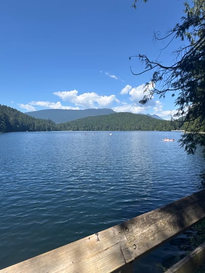 The image shows a scenic view of a lake in Port Moody, Canada, on a sunny day. The lake is surrounded by lush green trees and rolling hills in the background, under a bright blue sky with scattered white clouds. In the foreground, a wooden railing runs diagonally across the bottom of the frame, partially obscuring the view of the water.
Several people are visible on the lake, engaging in recreational activities. Two individuals are standing on paddleboards, while others are relaxing on a pink inflatable raft. A small white boat can also be seen in the distance. The water's surface is rippled, indicating a gentle breeze. The overall scene depicts a tranquil and inviting outdoor setting perfect for summer recreation.