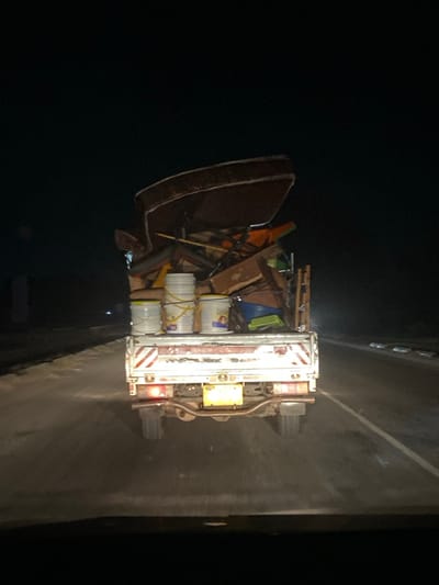 The image captures the rear view of a heavily loaded utility or pick-up truck at night, likely on a road in Bagamoyo, Tanzania. The scene is illuminated only by the truck's taillights and faint ambient light, emphasizing the darkness of the surroundings.
The truck, predominantly white, features vertical red and white striped reflective markings on its rear, common for commercial or utility vehicles to enhance visibility. Its open bed is packed with a large and varied collection of items. A prominent dark brown or maroon mattress leans against the left side of the cargo, slightly overhanging the truck's body. On the right side of the bed, several white plastic buckets, possibly containing paint or construction materials, are stacked. Amidst these, a chaotic mix of other objects is visible, including wooden planks, a long pole or broom, and various pieces of what appear to be furniture or general debris. The manner of loading suggests the items might be household goods being relocated or discarded.
No individuals are visible in the frame. The time of day is unmistakably night, evidenced by the deep darkness enveloping the scene and the illumination provided primarily by the truck's red taillights. Faint lights from distant vehicles or structures can be seen in the background on the road. The road surface appears to be asphalt and dry, with no specific weather conditions discernible. While there are some unidentifiable markings on the truck and buckets, no clear text is readable, nor is the license plate decipherable. The overall activity depicted is the nighttime transport of a bulky, possibly unsecured, load.