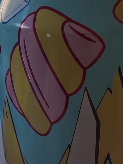 The image shows a close-up of a colorful illustration on a teal background. The illustration features rounded, abstract shapes in yellow and pink, outlined in dark red, resembling a stylized shell or a layered confection. In the lower right portion of the image, sharp, angular shapes in yellow and gray with dark outlines suggest a landscape or abstract mountain-like forms. The lighting suggests this is an indoor photograph, possibly of a printed material or artwork. The provided location of Дубрава-2 Снт, Russia is a rural settlement and does not provide specific visual context for the image itself.