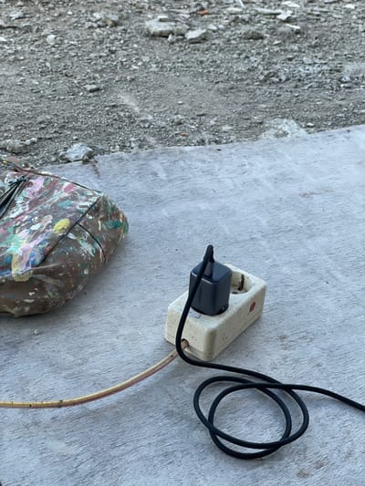The image displays an outdoor or semi-outdoor setting on a light grey, textured surface, possibly concrete, located in Mns Asan, Indonesia. An off-white, two-socket electrical power strip is positioned in the lower-middle right. A dark grey UGREEN brand wall charger is plugged into the left socket, from which a coiled black braided cable extends toward the lower right. A separate, yellowed electrical cable, marked with faint red spots, protrudes from the right side of the power strip, extending toward the lower left. To the immediate left, a brown canvas bag is visible, heavily stained with multiple colors of paint including pink, yellow, green, blue, white, and orange, with a zipper closure visible at its top edge. The upper half of the frame reveals a rough, unpaved ground surface consisting of grey gravel, small stones, and debris. No individuals are present in the scene.
