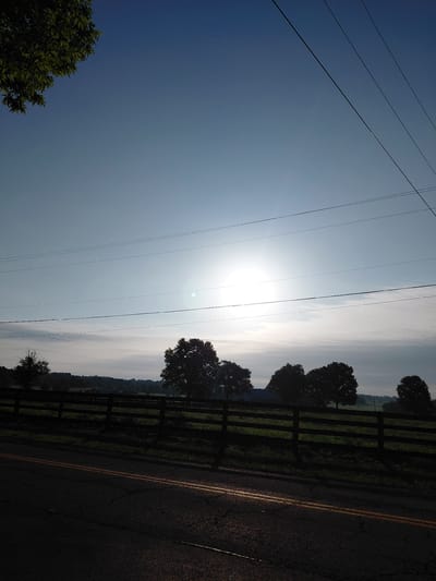 The image depicts a rural roadside scene at sunrise in Paris, United States. The foreground is dominated by a dark asphalt road with visible cracks and yellow dividing lines. To the left of the road, a dark wooden fence runs along a green grassy field. Several silhouetted trees are visible behind the fence, interspersed with the landscape. In the sky, several power lines stretch across the frame.
The sun is low on the horizon, casting a bright glare that obscures some of the details in the sky. The sky itself transitions from a deep blue on the left to a lighter, hazy white on the right, suggesting the early morning light. The overall impression is one of a tranquil rural morning. There is no text visible in the image.