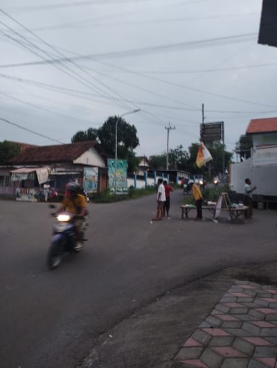 The image depicts an outdoor street scene, likely an intersection or junction, under an overcast or cloudy sky. The overall lighting suggests either early morning, late afternoon, or a very cloudy day.
In the foreground, a blue motorcycle with its headlight on is moving from right to left, with the rider visible in a yellow shirt and helmet, appearing blurred due to motion. To the right of the motorcycle, a patterned pavement made of interlocking red and grey blocks is visible.
Several people are gathered on the right side of the road, near a makeshift street vendor's stand made of wood. One person in a white shirt and dark pants stands with their back largely to the camera, facing another person in a red shirt. A third person in a yellow shirt is partially visible near the stand. Further back, another individual in a light-colored shirt is also present. They appear to be engaged in a social interaction or purchasing items from the stand. Small white specks, possibly litter, are scattered on the road.
In the mid-ground and background, buildings with reddish-brown tiled roofs and light-colored walls line the street. On the far left, a building has a small, blurry sign that appears to read "JUGLE" or "JUGGL" below its roofline. Numerous overhead power lines and utility poles crisscross the sky. Trees are scattered throughout the background.
A yellow and white flag or banner hangs from a pole in the middle-right background. Directly behind it, a dark signpost with white text is visible. The text clearly indicates street names: "JL. ANGGREK", "JL. FLAMBOYAN", "JL. MELATI", and also "WARUNG KUNING". These names strongly suggest the location is in Indonesia.
