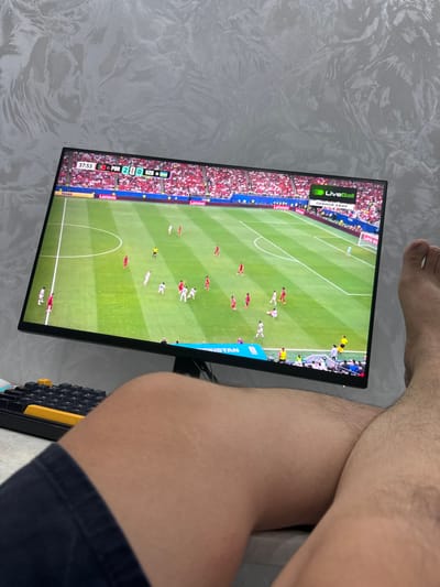 This is a medium shot of a person sitting and watching a soccer match on a monitor in Osh City, Kyrgyzstan. The monitor displays a live broadcast of a soccer game, with "LiveBall" and "ПРЯМОЙ ЭФИР" (which translates to "Live Broadcast" in Russian) visible at the top left. The score graphic at the bottom indicates Portugal (POR) leading Uzbekistan (UZB) 2-0 at 37:53 into the match. The setting appears to be indoors, possibly a bedroom, with a textured gray wall behind the monitor and the person's legs visible in the foreground. The person is wearing dark shorts. A keyboard with yellow and black keys is visible to the right of the monitor. The lighting suggests it could be daytime or the room is well-lit. The scene is focused on the person's engagement with the soccer game on the screen.