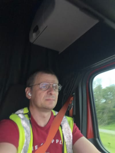 A middle-aged Caucasian male, wearing glasses and a red shirt with a high-visibility vest over it, is seen driving a vehicle. He has an earbud in his left ear and is wearing a seatbelt. The interior of the vehicle, including the dashboard and window, is visible. Outside the window, trees and a roadway can be seen. The image appears to be taken from inside the vehicle, looking towards the driver. There is no information or visual evidence to indicate the location in the city of Hillsboro, United States.