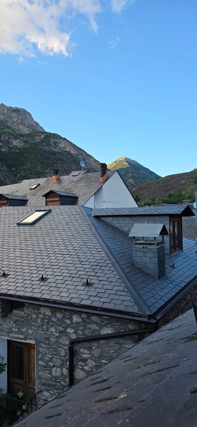 The image captures a scenic view from above the rooftops of traditional buildings, looking towards a majestic mountain range under a clear sky. In the foreground, several buildings are prominent, characterized by dark, interlocking slate tile roofs. These roofs feature brown rain gutters, multiple roof windows or skylights, and terracotta-colored chimneys. One roof also hosts a satellite dish and a larger structure that appears to be an outdoor cooking or ventilation unit. The walls of these buildings are constructed from rugged, irregularly shaped stones, giving them a rustic appearance.
The middle ground is dominated by a grand mountainous landscape, with steep, rocky peaks and slopes interspersed with green vegetation. The lighting on the mountains suggests either late afternoon or early morning, with parts of the peaks illuminated by sunlight and others cast in shadow. The upper portion of the image is filled with a vibrant blue sky, dotted with a few wispy white clouds, indicating clear and pleasant weather.
No people or explicit activities are depicted in the scene. The architecture and natural setting are consistent with a mountainous village environment, such as Benasque, Spain. There is no visible text in the image. The overall impression is one of tranquility and traditional mountain living.