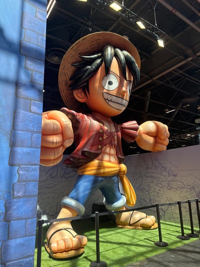 A large inflatable statue of the anime character Monkey D. Luffy stands on a patch of artificial grass in a convention hall. The statue is dressed in his iconic red shirt, blue pants, and straw hat. Behind the statue is a wall decorated with a world map. Black stanchions and ropes are set up in front of the statue, suggesting it is an exhibit. The ceiling of the hall is visible in the background, showing exposed beams, lighting fixtures, and ventilation systems. The statue is positioned near a blue, brick-patterned backdrop. This scene is located at the Paris-Nord Villepinte Exhibition Centre in Villepinte, France.