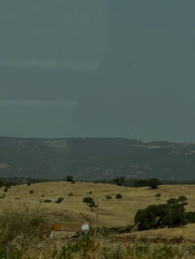 This is an image of a rural landscape. In the background, a dark, cloudy sky looms over a range of rolling hills covered in trees and vegetation. Several wind turbines are visible on the crest of the hills, with their white blades contrasting against the dark sky. The middle ground features a dry, grassy field dotted with scattered trees and bushes. A path or track winds through the field. In the foreground, there is a small, rustic building with a red-tiled roof and stone walls. Adjacent to the building is a white water tank or silo. The ground in the foreground is covered with dry grass, weeds, and some blurred foliage, suggesting movement or a slight blur from the camera. The overall impression is one of a remote, agricultural area with modern energy infrastructure. The location is Oschiri, Sardinia, Italy.
