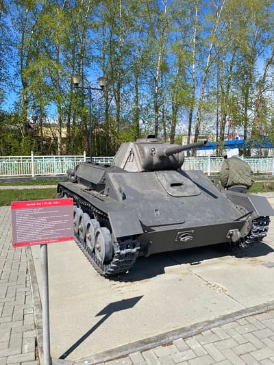 The image shows a T-70 light tank, specifically a model from 1942, on display in Kirovgrad, Russia. The tank is painted in a dark color, possibly gray or green. It is positioned on a paved area, likely a sidewalk or plaza. Behind the tank are trees with light green leaves against a blue sky. A white metal fence separates the tank from a grassy area. A sign next to the tank provides information about the T-70 in Russian, listing its specifications such as the year of production, crew size, dimensions, armament, armor thickness, engine power, speed, and obstacle-crossing capabilities. The sign also includes a warning not to go behind the fence. A shadow is visible on the pavement next to the tank, suggesting direct sunlight. A person is visible in the distance.