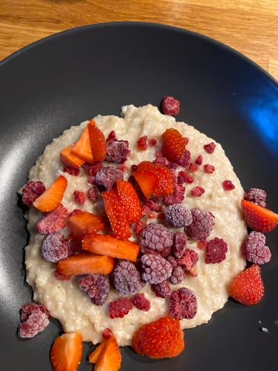 A top-down shot shows a black plate resting on a wooden surface. On the plate, there is a serving of what appears to be porridge or oatmeal. The dish is garnished with a scattering of fresh strawberries, some cut into slices and wedges, and a collection of frozen raspberries. The scene does not allow for location identification.