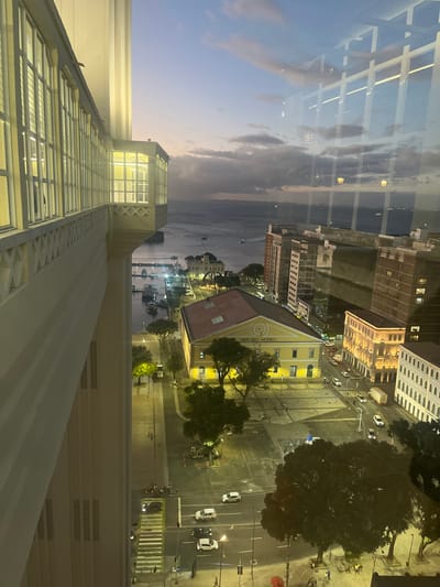 The image provides an elevated, dusk-time view of the waterfront in Salvador, Bahia, Brazil, specifically overlooking the Mercado Modelo and the Baía de Todos-os-Santos. The perspective appears to be from a building adjacent to or part of the Lacerda Elevator complex.
In the immediate foreground on the left, a portion of a cream-colored, multi-story building is visible, featuring a long horizontal section with numerous windows and an illuminated enclosed balcony area projecting outward, supported by a decorative railing. The building's interior lights are active.
The midground features the distinct yellow-painted Mercado Modelo building, with a dark red-brown gabled roof and a central circular facade displaying "MERCADO MODELO." This building is well-lit, casting light onto the surrounding area. In front of the Mercado Modelo is a large paved public square, mostly empty, with several dark green trees under artificial illumination.
Beyond the square, a multi-lane street carries vehicular traffic, including several white and dark cars, and a light-colored truck. Streetlights illuminate the roadway. A zebra crosswalk is visible in the lower-left, where a small group of indistinct figures can be seen.
Further into the midground, the Baía de Todos-os-Santos stretches across the scene. A pier with multiple small boats docked and moving is visible on the water's edge. Distant landmasses or structures are discernible across the bay under a twilight sky.
The background consists of the bay merging with a horizon line under a sky transitioning from pale blue at the horizon to a darker blue-gray overhead, with scattered dark clouds. Artificial lights are visible across the distant bay and on the landmasses.
To the right of the Mercado Modelo, various multi-story urban buildings with illuminated windows contribute to the city lights. Reflections of internal lights from the vantage point are visible on the glass through which the photograph was taken, particularly in the upper-right quadrant. The overall scene depicts an active urban environment transitioning into night, characterized by a mix of architectural structures, artificial illumination, and natural features.