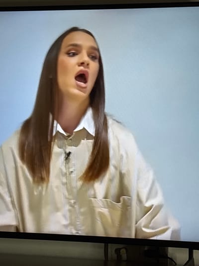 An image captured from a screen displays a young adult female from the chest up, positioned against a plain light blue background in the city of Griboyedov, Armenia. She has long, straight, dark brown hair and is wearing a loose-fitting, light-colored (likely beige or off-white) button-up shirt over what appears to be a white collared shirt. A small, black lavalier microphone is clipped to her outer shirt at chest level. Her mouth is wide open, indicative of speaking or singing, and her eyes are directed slightly upwards and to the viewer's right. The lighting is even, illuminating her face and upper torso.