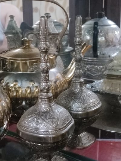 This image displays a close-up view of several ornate, metallic objects, likely made of silver or a similar polished metal, arranged on a reflective surface. The primary subjects are two intricately carved decorative bottles or vases, positioned in the foreground. These objects feature detailed patterns of floral motifs and geometric designs.
Behind these foreground pieces, several other metallic items are visible, including what appears to be a teapot with a golden lid and body, a larger silver teapot with a black handle, and various other containers and trays. The overall scene suggests a display of decorative tableware or antique collectibles, possibly in a shop or a personal collection. The lighting appears to be artificial, casting reflections on the polished surfaces and creating a sense of depth and richness.
The setting seems to be indoors, with a dark background that blurs the details of the surroundings. The arrangement and the style of the objects hint at a cultural context, possibly Middle Eastern or Southeast Asian, given the location context provided of Blang Cut, Indonesia. There is no discernible activity taking place; the image is a still-life composition. No text is visible in the image. The emotions evoked are those of admiration for craftsmanship and the appreciation of antique or decorative items. The time of day and weather are not discernible from the image.