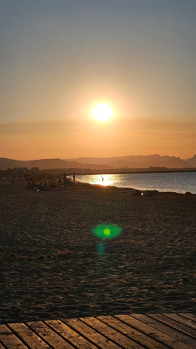 This is a vertical, eye-level, full shot of a beach at sunset in Deveses, Spain. The sun is low in the sky, casting a warm, orange glow over the scene. The sun is a bright, white orb in the upper center of the image. The sky above the horizon is a gradient of light blue to pale orange, transitioning to a deeper orange closer to the sun. The ocean is calm and reflects the sun's light, creating a shimmering path across the water.
In the middle ground, a sandy beach is populated with several groups of people. Some are sitting or lying on the sand, while others are standing or walking. Several individuals are in the shallow water, with a few people swimming. The coastline stretches into the distance, with hazy, layered mountains forming a backdrop. Buildings are visible along the distant shore.
The foreground is dominated by the textured surface of the sandy beach. In the lower right corner, a portion of a wooden boardwalk or pier is visible, with planks running diagonally. A green lens flare is present in the sand in the mid-ground, slightly to the left of the center. The overall atmosphere is tranquil and serene, capturing the beauty of a sunset at the coast.