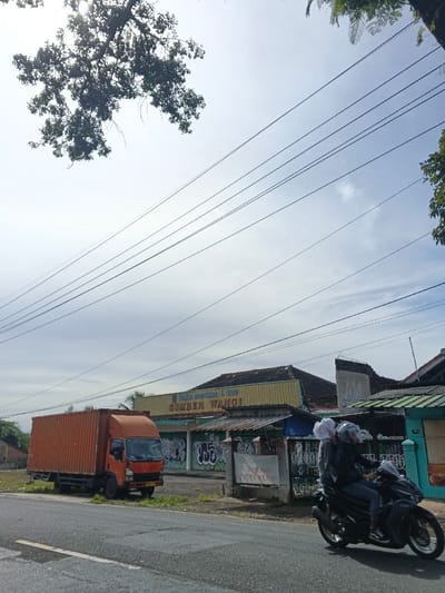 The image captures a bright, daytime street scene, likely in a semi-urban or rural area, consistent with the Carcar, Philippines context. The sky is bright with scattered clouds, and numerous power lines stretch diagonally across the upper and middle portions of the frame. Silhouetted tree branches with dense foliage are visible in the upper left, and lighter green leaves appear in the upper right.
In the mid-ground, an orange box truck is parked on a dirt shoulder next to the asphalt road. Behind it is a low-rise building with a yellowish-green facade and a corrugated metal roof. A sign on this building reads "TOKO ROTI MANISAN & KUE" (Sweet Bread & Cake Shop) and "SUMBER WANGI" (Fragrant Source), which appears to be Indonesian. Below the sign, the building wall is covered in graffiti. To the right of this building, other residential or commercial structures are visible, some with green-colored roofs or exterior elements.
On the road in the foreground, a dark-colored scooter or motorcycle with two riders is in motion, heading right. The driver wears a dark jacket and a helmet, while the passenger, also wearing a dark jacket, has a white head covering. The road surface is asphalt, with some patches and lines visible. The overall impression is a bustling roadside environment under a clear sky.