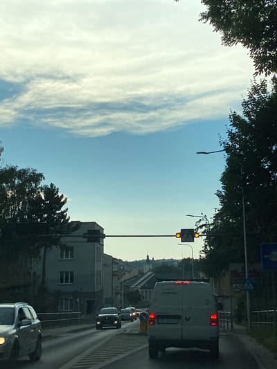 The image depicts a street scene in Bielsko-Biała, Poland. The perspective appears to be from inside a vehicle, looking forward and slightly upwards. The sky is a clear blue with wispy white clouds, suggesting it is daytime, likely late afternoon or early evening due to the soft lighting. Trees with dark green foliage frame the top of the image, casting some shade.
In the foreground, there are several vehicles on a paved road. The closest vehicle is a silver car, seen from the side. Further ahead, a dark-colored car is visible, followed by a white van. The van has its brake lights illuminated, indicating it may be slowing down or stopped. The license plate of the van is partially visible, showing "SB 353PH". Text on the side of the van reads "MEINHOFER".
Traffic signals are visible, with one showing two orange lights, suggesting a caution or impending change. A directional sign with an arrow pointing left is also present. Buildings line the right side of the street, appearing to be residential or commercial structures. In the distance, a cityscape with buildings and a church spire is visible against the horizon. The overall atmosphere is calm and mundane, capturing a typical urban environment during daylight hours.