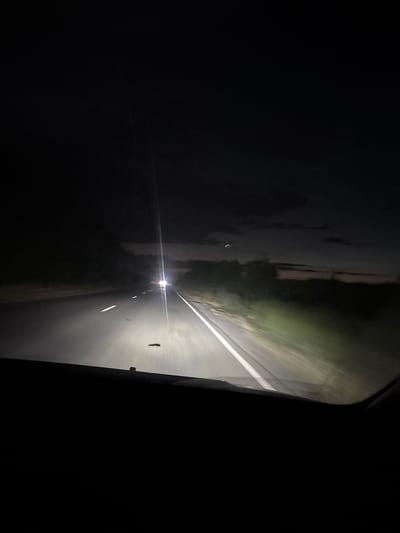The image is a first-person perspective, taken from inside a vehicle at night. The vehicle is on a paved road with white lane markings. The headlights of the vehicle illuminate the road ahead, creating a bright white glow that stretches into the darkness. On the right side of the road, the headlights also illuminate a blurry embankment of vegetation. In the distance, the headlights of an oncoming vehicle are visible, appearing as a bright light source with a strong glare. The sky above is dark, with some faint clouds visible, and there is a sense of a twilight or dusk transitioning into night. The overall scene depicts driving on a rural road at night. The location is stated to be in the city of Hatsik, Armenia.