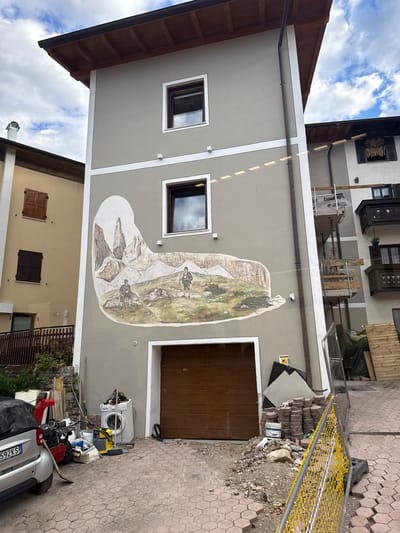 The image shows a multi-story building in Cavedago, Italy, with a large mural painted on its side. The mural depicts a mountainous landscape with two figures dressed in hiking attire. The building has several windows and a brown garage door at ground level. In front of the building, there are various objects, including a car, a washing machine, gardening tools, and construction materials like bricks and wooden planks. A yellow safety fence is also visible. The sky above is partly cloudy. The building appears to be a residential or commercial property, and the surrounding area seems to be under some form of construction or renovation.