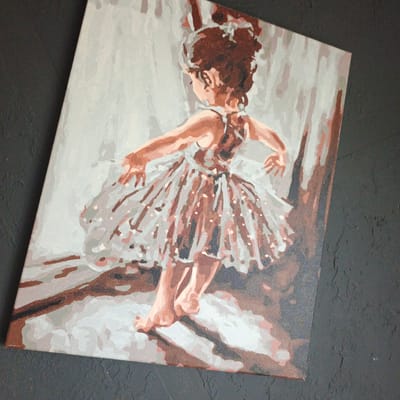 A painting on canvas depicts a young ballerina in a tutu, seen from the back. The ballerina has her arms outstretched and is on pointe. The background of the painting appears to be a light gray, with some vertical elements that could represent curtains or a stage backdrop. The overall style is impressionistic, with broad brushstrokes and a limited color palette dominated by browns, grays, and whites. The painting is placed on a dark, textured surface, possibly a wall or table, which is out of focus. There is no indication of the city of Vyshgorodok, Russia, or any other specific location in the image.