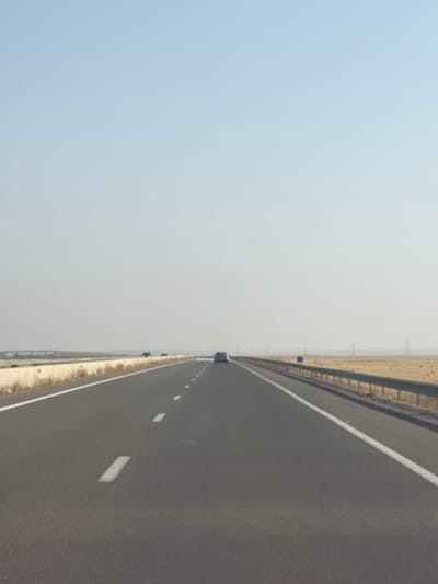 The image shows a highway in Nzalat Laadam, Morocco. The highway has a dark gray asphalt surface with white lane markings. A white line marks the edge of the road. To the right of the highway is a sandy area with sparse vegetation. A gray sky is visible on the left side of the image. A vehicle is seen in the distance.