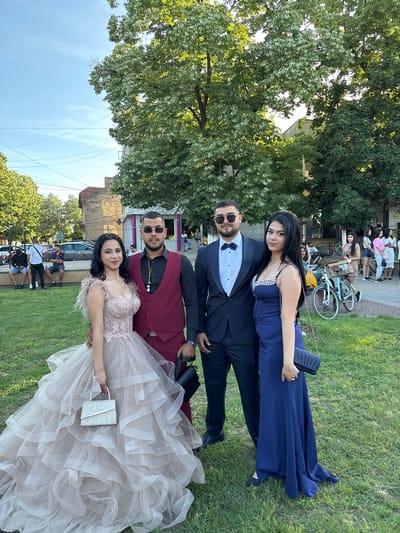 Two couples pose on a grassy area in Hayredin, Bulgaria. The first couple features a woman in a long, dark blue dress with a beaded bodice, holding a dark clutch. She stands next to a man in a black tuxedo with a bow tie and sunglasses. The second couple consists of a woman in a light-colored, frilly gown holding a silver clutch. She stands next to a man in a maroon vest, black shirt, and sunglasses. In the background, there are people, bicycles, and buildings. Trees are also visible in the background, and the sky is partly cloudy.